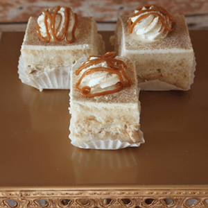 Three AP-045M Apple Cinnamon Chai Squares, featuring creamy filling and topped with whipped cream and caramel drizzle, are presented on a gold stand with crystal accents in white paper cups.