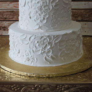 Product WD-005: A two-tier white cake with elegant floral designs in white icing, presented on a gold-embossed stand against a rustic wooden backdrop.