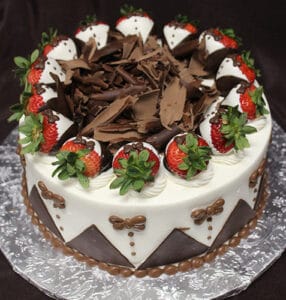 BD-019 is a round cake topped with chocolate-dipped strawberries and chocolate shavings, covered in white icing with chocolate designs on the sides, and presented on a silver foil-covered board.
