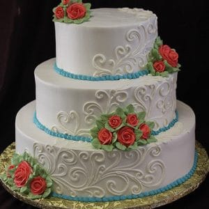 The WC-046 is a three-tiered white cake featuring intricate swirl designs, blue bead borders, clusters of red and pink frosting roses with green leaves, all elegantly presented on a gold platter.