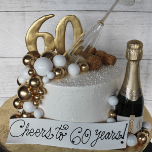 The BD-024U Celebration cake is a white frosted cake on a gold board, decorated with gold and white spheres, a gold "60" topper, tipped wine glass, corks, mini champagne bottle, and a "Cheers to 60 years!" sign.