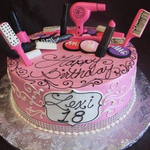 The BD-028B Makeup/Hair is a pink birthday cake with fondant makeup and hair styling decorations, white swirls, a “Lexi 18” plaque, and “Happy Birthday” elegantly scripted on top.