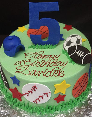 CB-017B is a round birthday cake decorated with sports-themed items, a blue cap, colorful stars, and a large blue number 5 on top. "Happy Birthday Davidek" is written in red icing on green frosting.