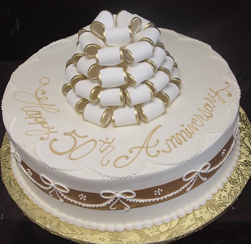 AN-020: A round white cake with “Happy 50th Anniversary” in gold icing, topped with white and gold ribbon loops, and finished with a brown ribbon and white bows around the sides.