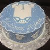 BB-049: Round cake with blue and white icing, baby onesie design on top, white polka dots, "Welcome Adam" text, and sides decorated with small white onesies.