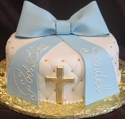 The RC-059 is a round white cake with a large blue fondant bow, gold accents, a gold cross on the side, and "God Bless Sendel" written in white script on the blue ribbons.