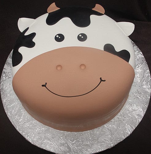 The CB-012C is a round cake designed as a cute cow's face, featuring black and white patches, small ears, nostrils, and a smiling mouth. It is presented on a silver floral-patterned cake board.