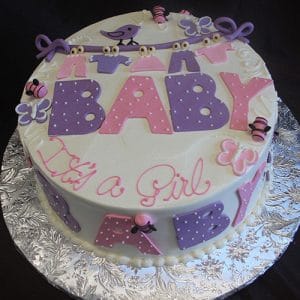 BB-017 is a round cake with pink and purple fondant “BABY” letters, polka dots, butterflies, bees, and a bird banner. “It’s a Girl” in pink script decorates the side. Presented on a silver floral-patterned board.