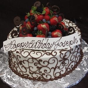 BD-162 is a round cake with white icing, chocolate swirls, assorted berries, and chocolate curls. A chocolate plaque reads "Happy Birthday Joseph." Presented on a silver tray.