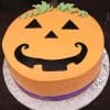 The HW-004 cake is round, decorated as a smiling jack-o-lantern with orange frosting, black eyes, nose, and mouth, green icing stem accents on top, and a purple band around the bottom edge.