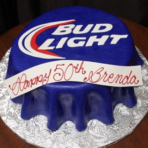 The BD-025 Bud Light is a round cake designed as a blue Bud Light can, featuring the logo on top and a white banner with "Happy 50th Brenda" in red script, set on a silver patterned board.