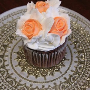 A chocolate cupcake named CC-013 Gold, filled with chocolate mousse and topped with white frosting and three peach-colored buttercream roses, elegantly displayed on an ornate silver plate.