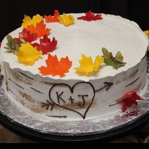 BR-051 is a round cake with white frosting, decorated with colorful autumn leaves and a heart carved with K + J to resemble a birch tree. It’s presented on a foil-covered board.