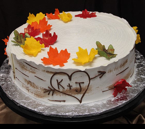 BR-051 is a round cake with white frosting, decorated with colorful autumn leaves and a heart carved with K + J to resemble a birch tree. It’s presented on a foil-covered board.