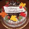 The THX-001 Display Strawberry Grand Marnier Cake with Fall Decor is a round cake adorned with chocolate shavings, dipped strawberries, colorful fondant leaves, and a white plaque reading "Happy Thanksgiving" in elegant script.
