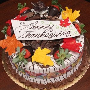 The THX-001 Display Strawberry Grand Marnier Cake with Fall Decor is a round cake adorned with chocolate shavings, dipped strawberries, colorful fondant leaves, and a white plaque reading "Happy Thanksgiving" in elegant script.