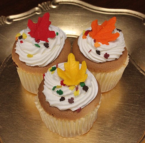 The CC-068 Display Fall Decor Gold cupcake features chocolate mousse filling, white frosting, colorful leaf-shaped sprinkles, and large red, orange, and yellow leaf decorations on top, served on a gold plate.