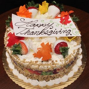 The THX-002 Display Carrot Cake with Fall Decor is a round cake topped with strawberries, white chocolate-dipped berries, colorful fondant leaves, and a “Happy Thanksgiving” card in script, served on a gold tray.