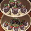 A two-tiered gold tray displays ST-000 Dark Chocolate Strawberries, each drizzled with white chocolate and set in a white paper cup, on a wooden surface.