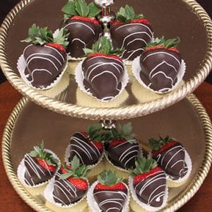 A two-tiered gold tray displays ST-000 Dark Chocolate Strawberries, each drizzled with white chocolate and set in a white paper cup, on a wooden surface.