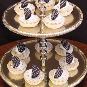 Two-tiered gold tray displaying Oreo Cheesecake mini tarts topped with whipped cream and a chocolate-dipped cookie piece, neatly arranged on both levels against a dark background.