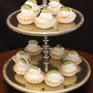 A two-tiered silver stand displays Keylime Tarts topped with whipped cream and lime slices. The dark background highlights the vibrant desserts.