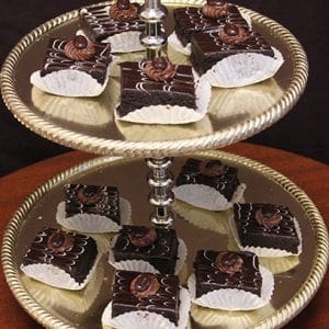A two-tiered gold dessert stand displays Chocolate Decadence Gluten Free pastries—chocolate-covered squares in white paper cups with a chocolate swirl on top—neatly arranged on both levels.