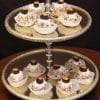 A two-tiered gold tray displays White Chocolate Chapeaux—cupcakes with nuts, white frosting, and chocolate or caramel pieces in white liners—arranged against a dark background.