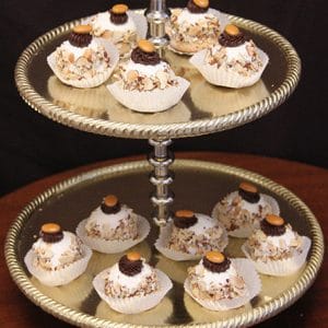 A two-tiered gold tray displays White Chocolate Chapeaux—cupcakes with nuts, white frosting, and chocolate or caramel pieces in white liners—arranged against a dark background.