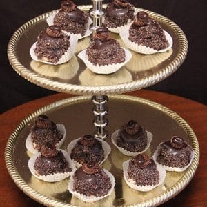 A two-tiered silver stand displays Chocolate Chapeaux truffles, each topped with a chocolate decoration and set in a white paper cup, resting on a dark brown surface against a black background.