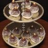 The AP-025 Hazelnut Chapeaux are displayed on a two-tier gold dessert stand, each chocolate truffle in a white paper cup, topped with hazelnuts and chocolate drizzle, set on wood against a dark background.