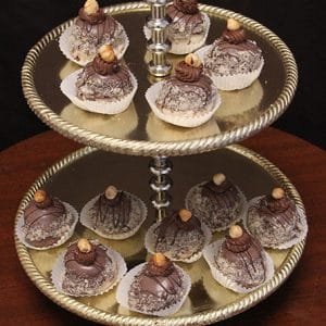 The AP-025 Hazelnut Chapeaux are displayed on a two-tier gold dessert stand, each chocolate truffle in a white paper cup, topped with hazelnuts and chocolate drizzle, set on wood against a dark background.