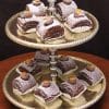 A two-tiered gold tray displays Chocolate Roulades, each square dusted with powdered sugar, topped with decorative chocolate and caramel pieces, and placed in white paper cups.