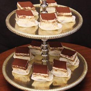 Tiramisu Diamonds, served in white paper cups and dusted with cocoa powder, are neatly arranged on both tiers of a silver serving tray against a dark background.