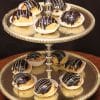 A two-tiered gold tray holds nine Ganache Cream Puffs, each topped with chocolate glaze and a white icing drizzle, placed in white paper cups.