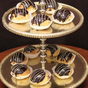 A two-tiered gold tray holds nine Ganache Cream Puffs, each topped with chocolate glaze and a white icing drizzle, placed in white paper cups.