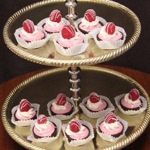 A two-tiered silver dessert stand displays twelve AP-032 Chocolate Raspberry Cups with pink frosting, each topped with a fresh raspberry and white chocolate drizzle, arranged neatly in white paper cups.