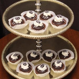 A two-tiered silver serving tray displays twelve Chocolate Cups in paper cups, each topped with white cream and drizzled chocolate, set against a dark background.