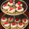 A two-tiered gold serving tray displays Mini Cheesecakes, each topped with whipped cream and fresh strawberries, arranged on both tiers against a dark background.