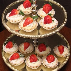 A two-tiered gold serving tray displays Mini Cheesecakes, each topped with whipped cream and fresh strawberries, arranged on both tiers against a dark background.