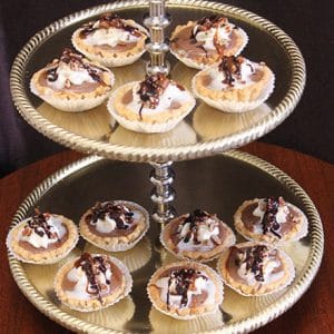A two-tiered serving tray displays Turtle Cheesecake slices topped with whipped cream and chocolate drizzle, arranged neatly on both levels against a dark background.