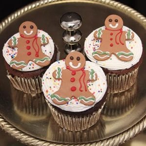 Three CH-066A gold cupcakes with chocolate mousse filling, topped with white frosting, colorful sprinkles, and smiling gingerbread men, displayed on a silver tray.