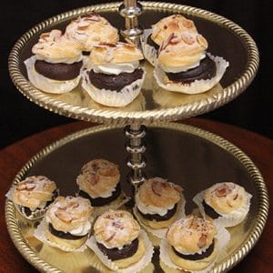 A two-tiered gold dessert stand displays Hazelnut Cream Puffs, each topped with sliced almonds and chocolate and placed in a paper wrapper, set against a dark background.