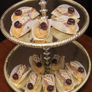 A two-tiered silver serving tray holds Chocolate Eclairs, each drizzled with chocolate, topped with a chocolate garnish, and placed in white paper cups. The eclairs are arranged neatly on both tiers of the tray.