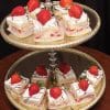 The gold two-tier dessert stand holds Strawberry Grand Marnier Squares—cake pieces topped with whipped cream, chocolate drizzle, and fresh strawberries. The trays have a decorative edge and are set against a dark background.