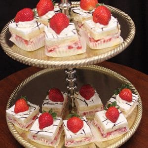 The gold two-tier dessert stand holds Strawberry Grand Marnier Squares—cake pieces topped with whipped cream, chocolate drizzle, and fresh strawberries. The trays have a decorative edge and are set against a dark background.