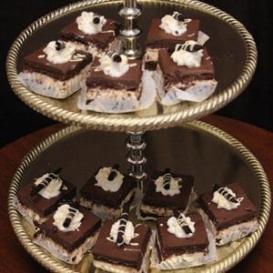 A two-tiered gold tray displays Peanut Butter Squares, each topped with a swirl of cream and chocolate garnish on white paper liners, set against a dark background.