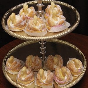 The Chantily Swan, cream puff pastries shaped like swans and dusted with powdered sugar, are arranged in paper cups on a two-tiered silver tray atop a dark wooden table against a black background.