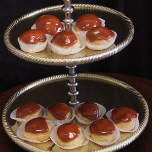 A two-tiered silver tray displays Caramel Cream Puffs, each in a white paper cup and topped with glossy caramel glaze. The dark background highlights the delicate treats.