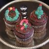 Three CC-077 Display Christmas Decor Chocolate cupcakes with white filling sit on a silver tray, each topped with colorful Christmas-themed icing—wreath, candy cane, and Christmas tree—and finished with sprinkles.